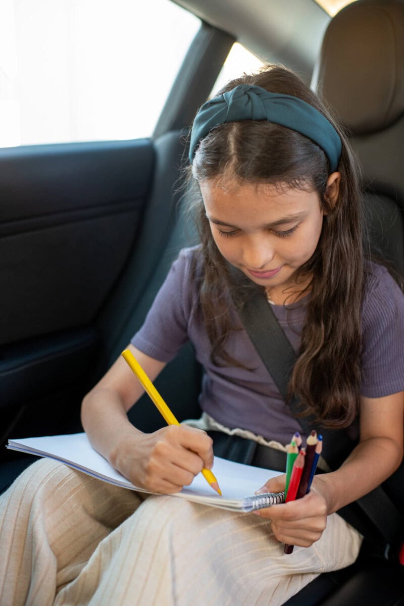 girl writing in a pad in the back seat of a car on a road trip