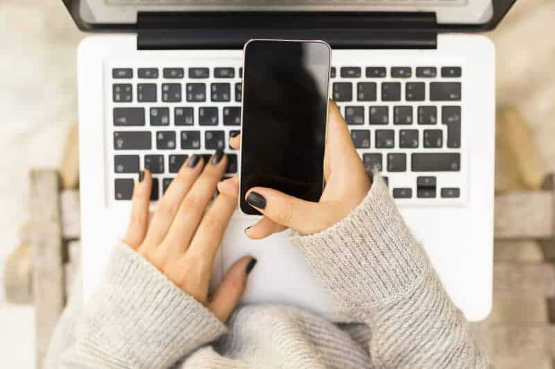 woman's hands holding a phone and typing on a laptop
