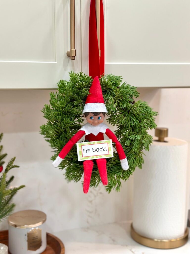 Elf sitting in a wreath with a sign that says "I'm back"