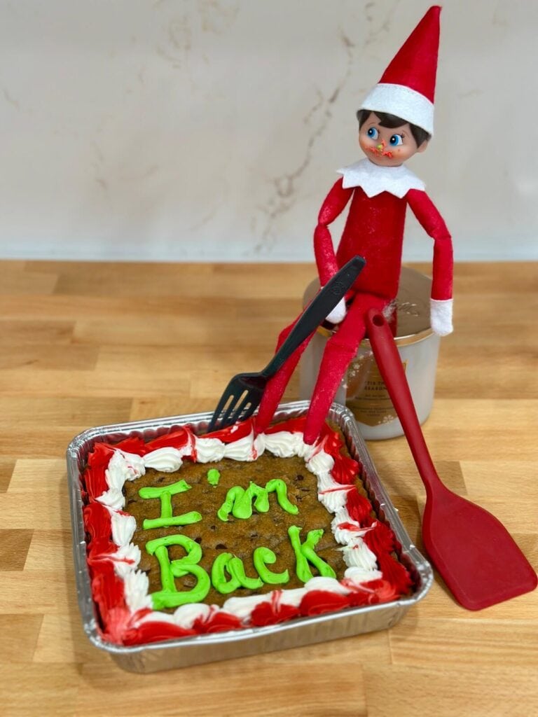 Elf on the Shelf sitting with a cookie cake that says "I'm back" in icing