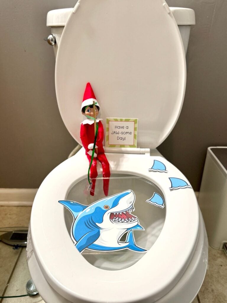 Elf on the Shelf sitting on a toilet seat with a picture of a shark in the toilet bowl