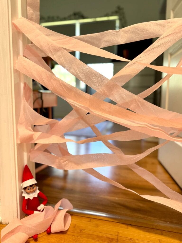 elf on the shelf sitting in front of a bedroom doorway with toilet paper taped across it